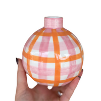 Thumbnail for A hand holds the Noss & Co Bud Vase Round - Pink & Orange Gingham, a stoneware porcelain vase with a narrow neck, featuring a pink and orange gingham design on white. The background is plain white.