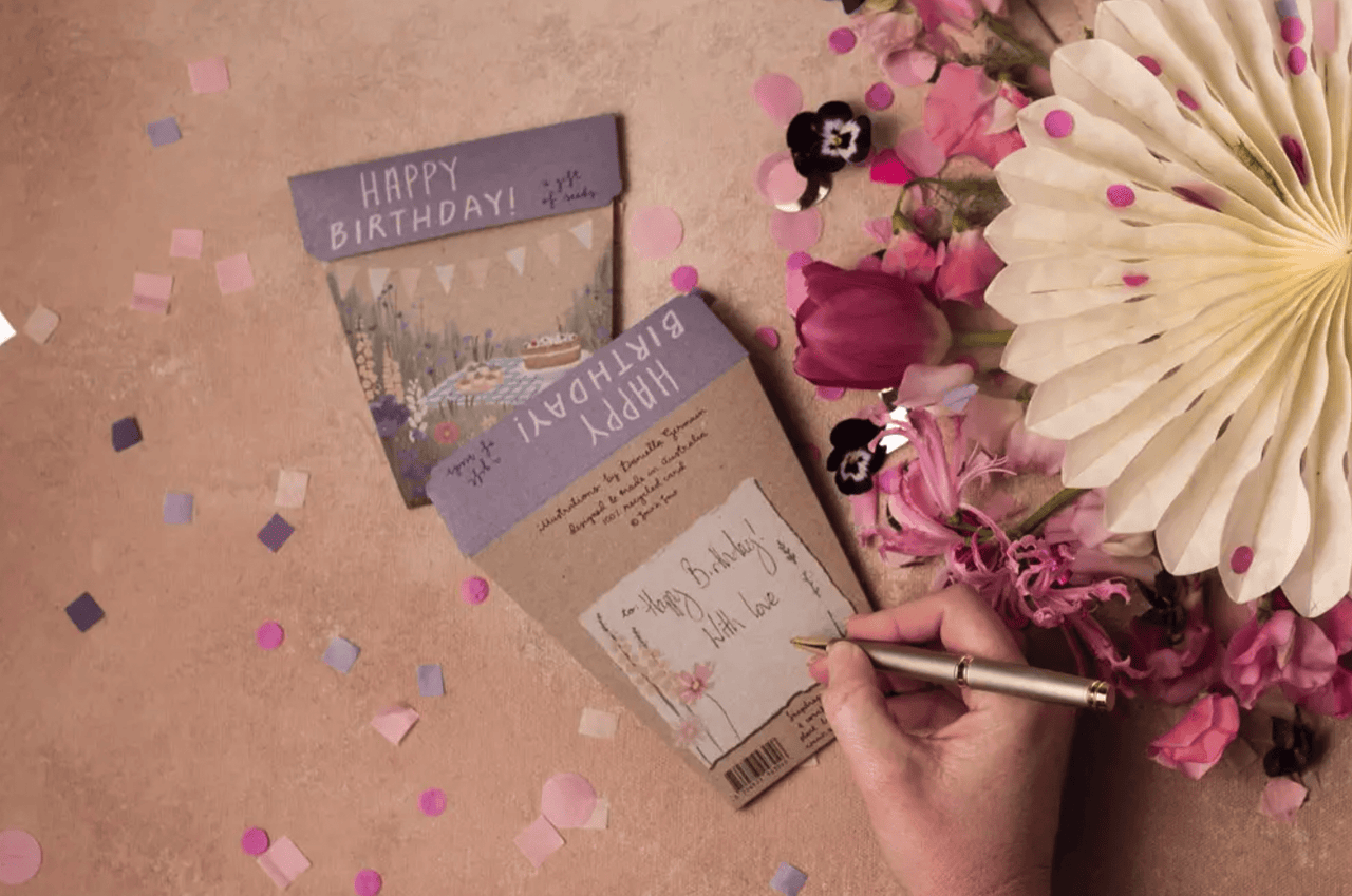 A person writing a birthday card in front of Sow n Sow's 'Happy Birthday' Picnic seeds.