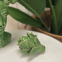 Thumbnail for A green head-shaped ceramic vase with plants sits beside a textured Artichoke - Green by Mediterranean Markets on a white surface, complemented by large green leaves in the background for a vibrant, natural ambiance.