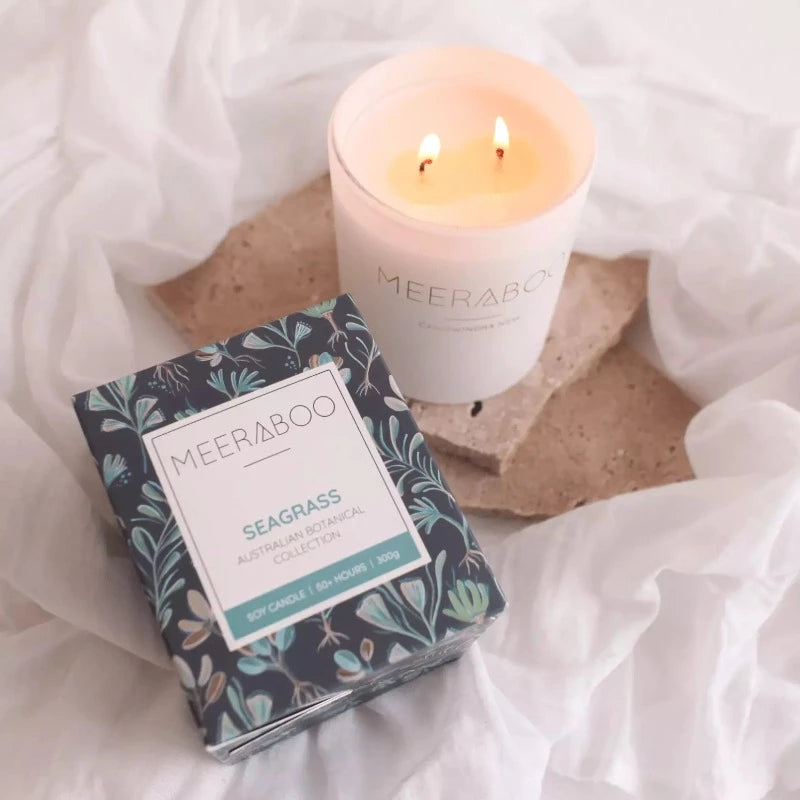 A Meeraboo Boxed Soy Candle - Seagrass 300g with a box next to it on a bed.