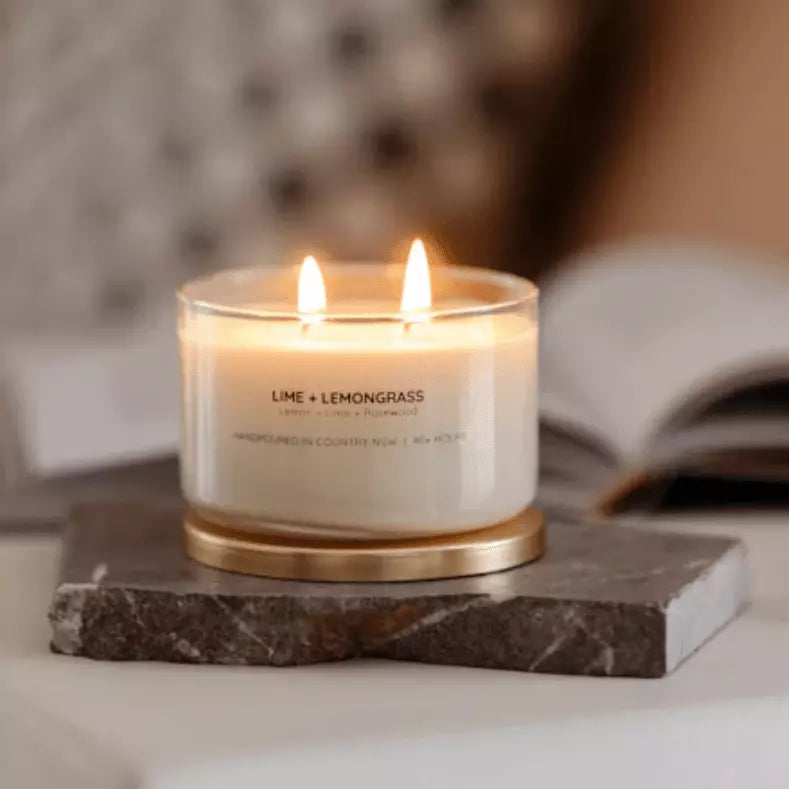 A Meeraboo Soy Candle - Lime + Lemongrass sits on a marble table next to a book.