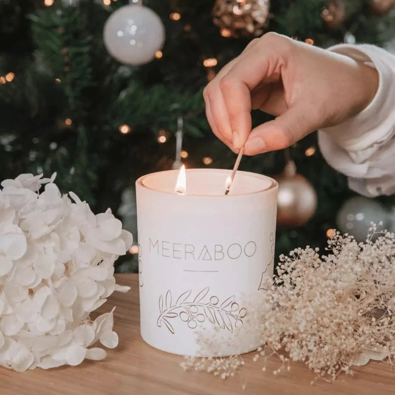 A person lighting a Meeraboo Boxed Soy Candle - Plum + Pomegranate in front of a christmas tree.