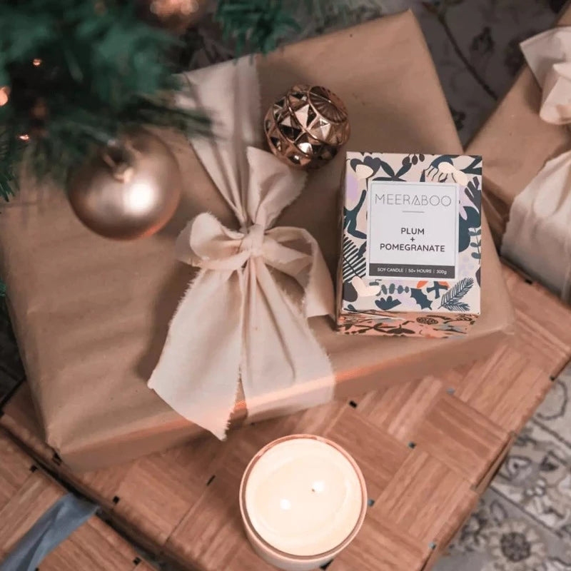 A Meeraboo boxed soy candle - Plum + Pomegranate and a Christmas tree on a wooden table.