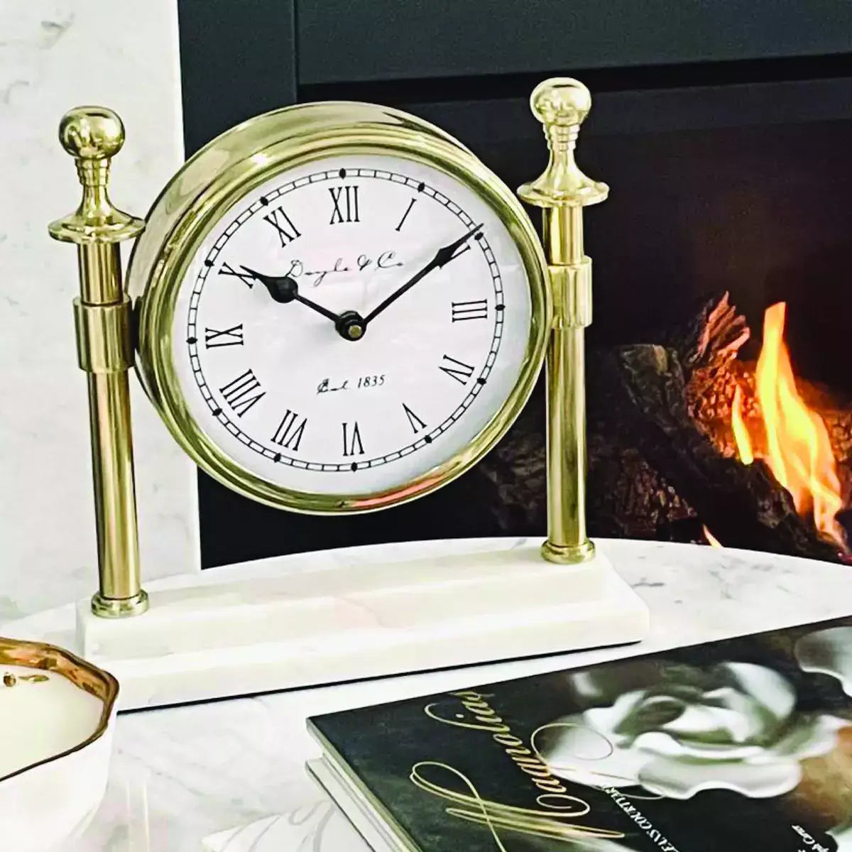 A Column Clock with White Marble Base - Gold sits on a table next to a fireplace.