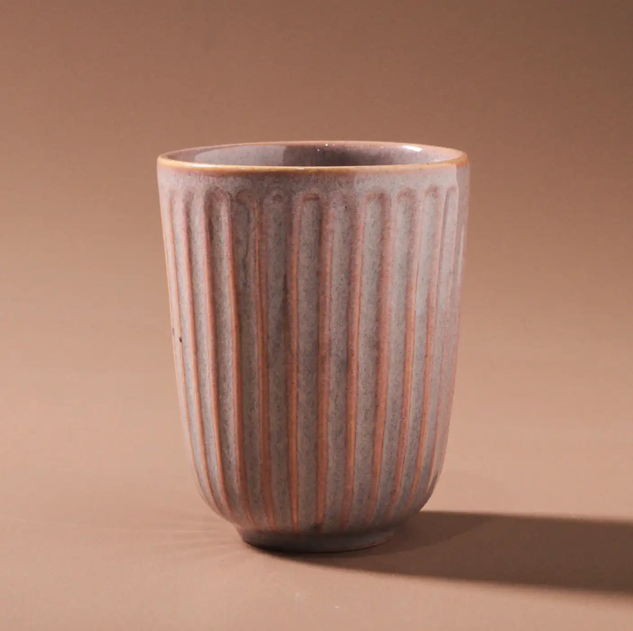 The Juliette Latte Mug - Pink by Indigo Love features a vertical ribbed texture and matte finish on a neutral brown backdrop. This ceramic mug flaunts muted earthy tones with subtle gradients, showcasing its handcrafted charm enhanced by reactive glaze elements.