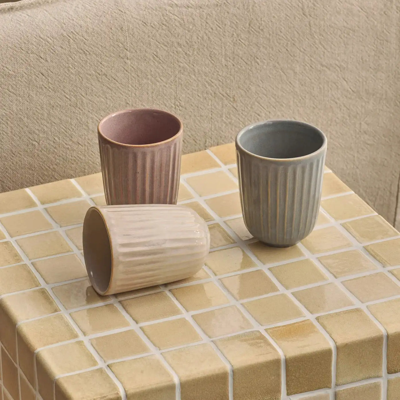 Three handcrafted Juliette Latte Mugs by Indigo Love, featuring vertical ridges, sit on a tiled surface with cream and light brown tiles. The mugs come in muted pink, beige, and light blue colors against a beige fabric backdrop.