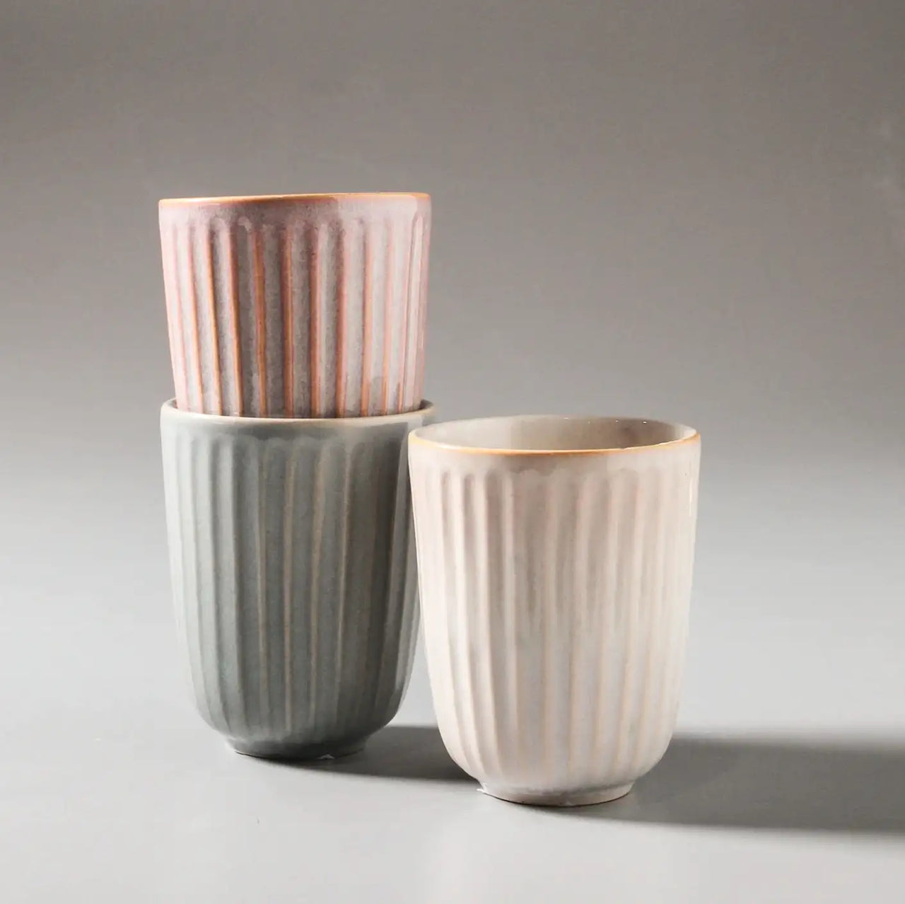 Three Juliette Latte Mugs from Indigo Love, in pastel shades, are stacked artfully: pink on top, gray in the middle, and off-white in the foreground against a soft gray background, highlighting their artisanal charm.