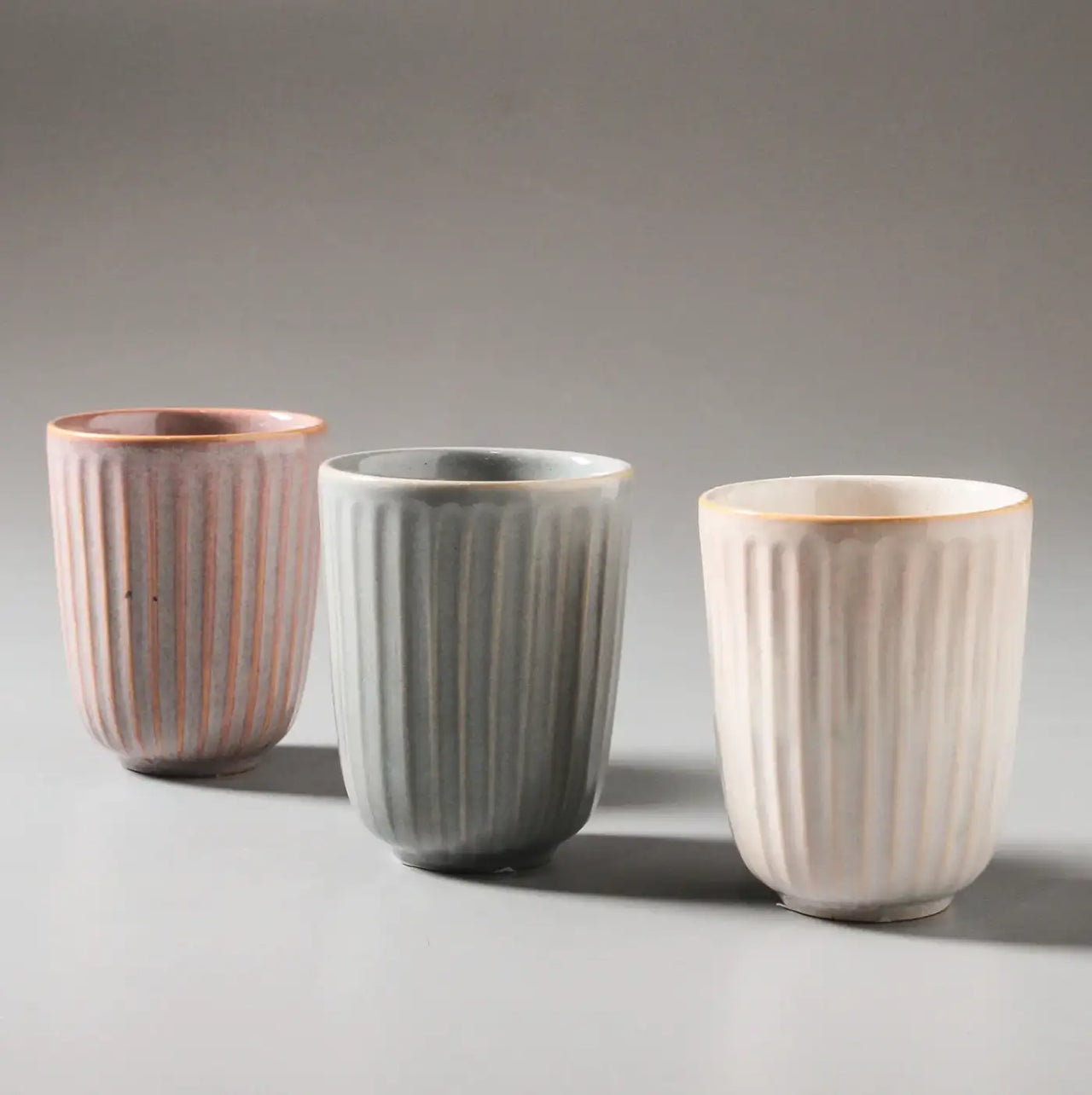 The Juliette Latte Mugs by Indigo Love, featuring a reactive glaze with vertical stripes, are arranged on a light gray surface in pastel pink, gray, and white for an elegant and minimalist display.