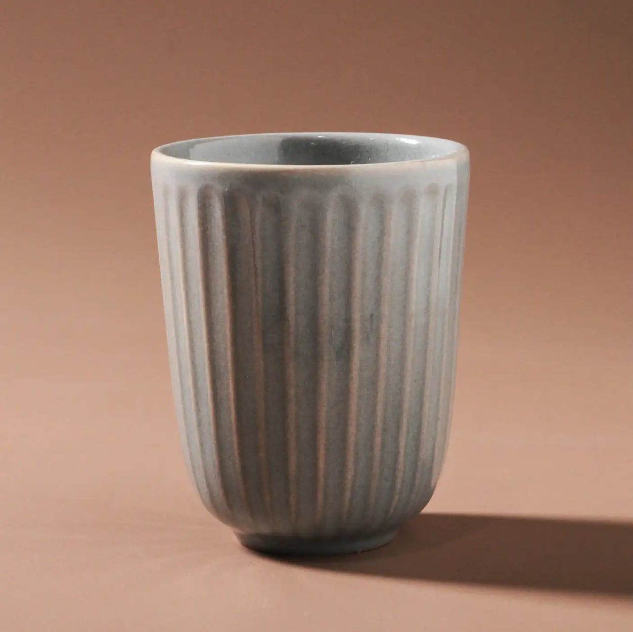 The Juliette Latte Mug by Indigo Love, in a sleek light blue with vertical texture, is centered against a soft brown background, casting a subtle shadow. Each unique, hand-finished ceramic piece adds elegant distinction to your collection.