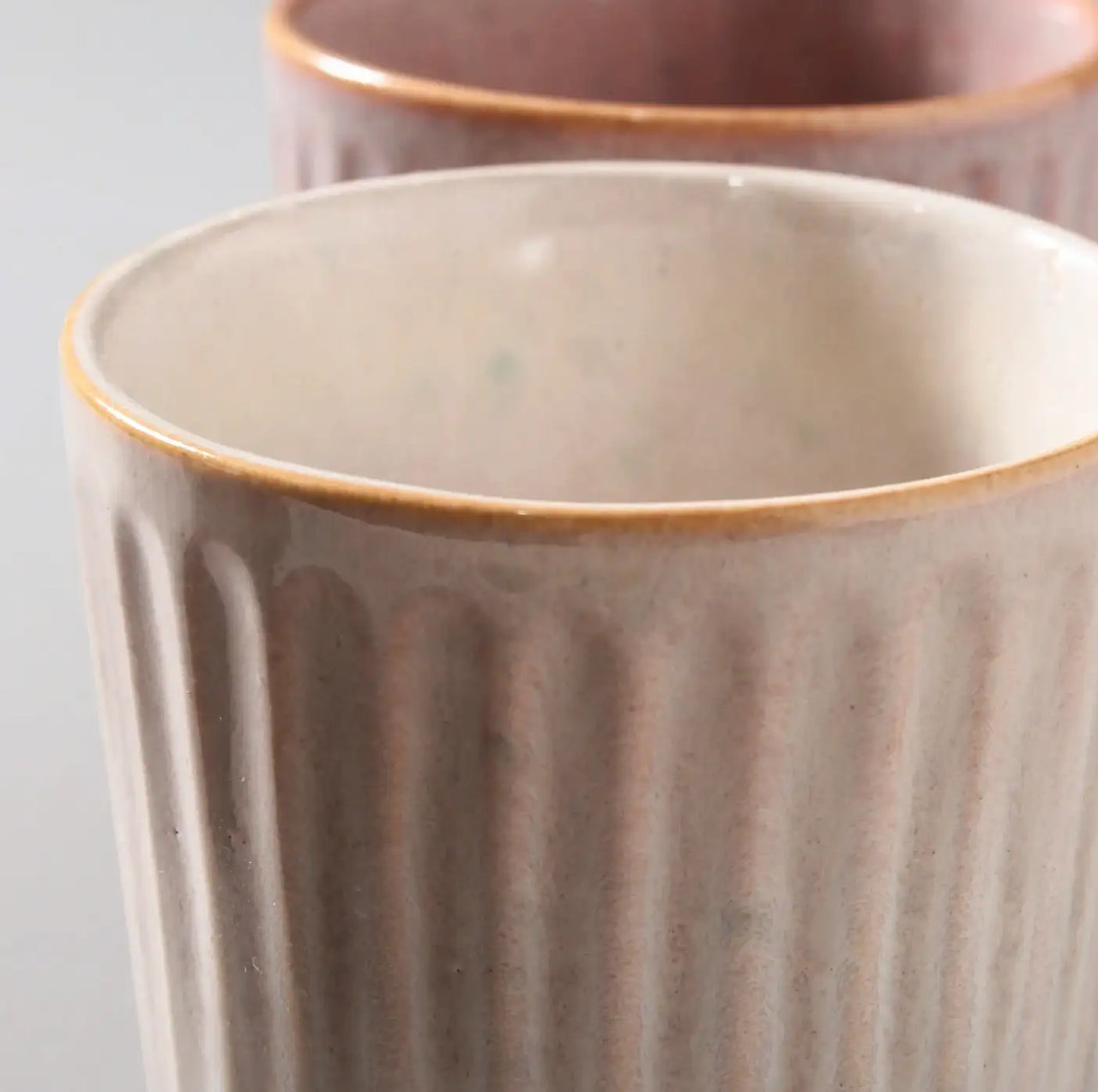 Close-up of two Juliette Latte Mugs by Indigo Love, showcasing hand-finished ceramic appeal. The front mug is cream with vertical grooves and a brown rim, while the partially visible background mug is pink, both featuring similar design elements.