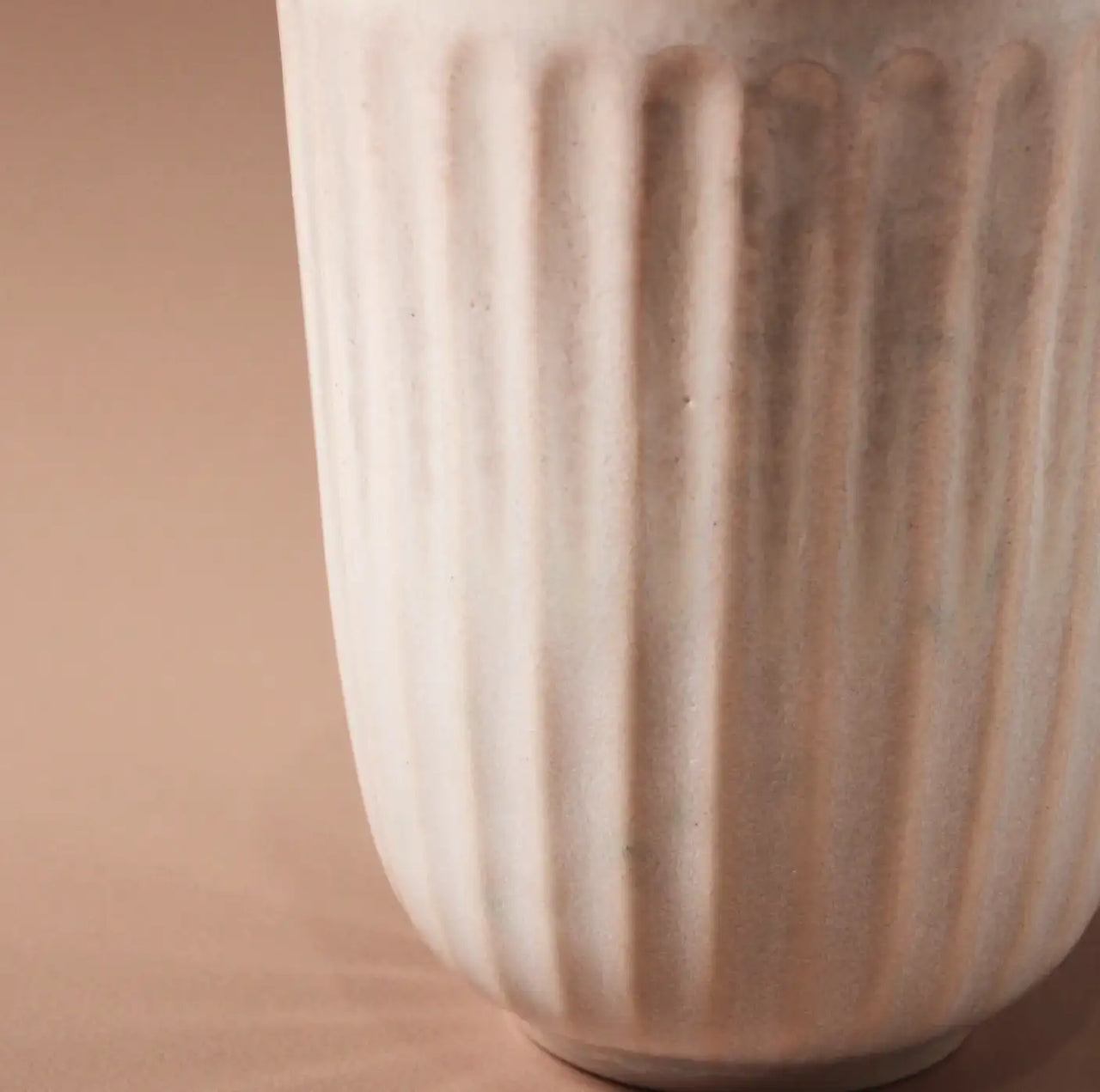 A close-up of a textured ceramic vase with vertical grooves on a beige background reveals a soft, matte finish and subtly tapered shape similar to the Juliette Latte Mug. Made by Indigo Love, the white mug embodies the elegance of hand-finished ceramics.