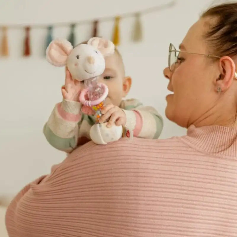 A baby is held on an adult’s shoulder, clutching the Pilbeam Fehn Rainmaker Mouse plush motor skills toy with a rattle. The adult, in glasses and a pink ribbed sweater, stands before colorful tassel decorations ideal for baby showers or sensory play.
