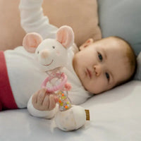 Thumbnail for A baby in a white top and red pants lies on a bed, holding the Pilbeam Fehn Rainmaker Mouse—a soft motor skills toy with pink ears and a smiling face—while looking at the camera.