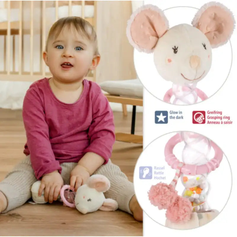 A toddler in a pink shirt and beige pants sits holding the Pilbeam Fehn Rainmaker Mouse, a plush motor skills toy with glow-in-the-dark details, grasping ring, and rattle—a perfect sensory gift for baby showers.