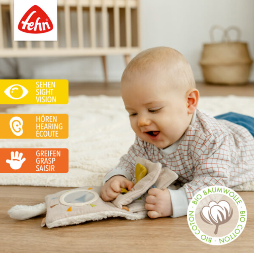 A baby plays on the floor with the Pilbeam FehnNATTUR Soft Book Sheep, a sensory activity toy made from organic cotton. Icons on the left highlight sight, hearing, and grasping features. A bio cotton label and wooden crib appear in the background.