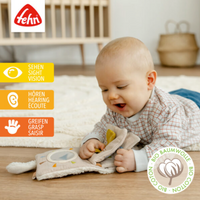 Thumbnail for A baby plays on the floor with the Pilbeam FehnNATTUR Soft Book Sheep, a sensory activity toy made from organic cotton. Icons on the left highlight sight, hearing, and grasping features. A bio cotton label and wooden crib appear in the background.