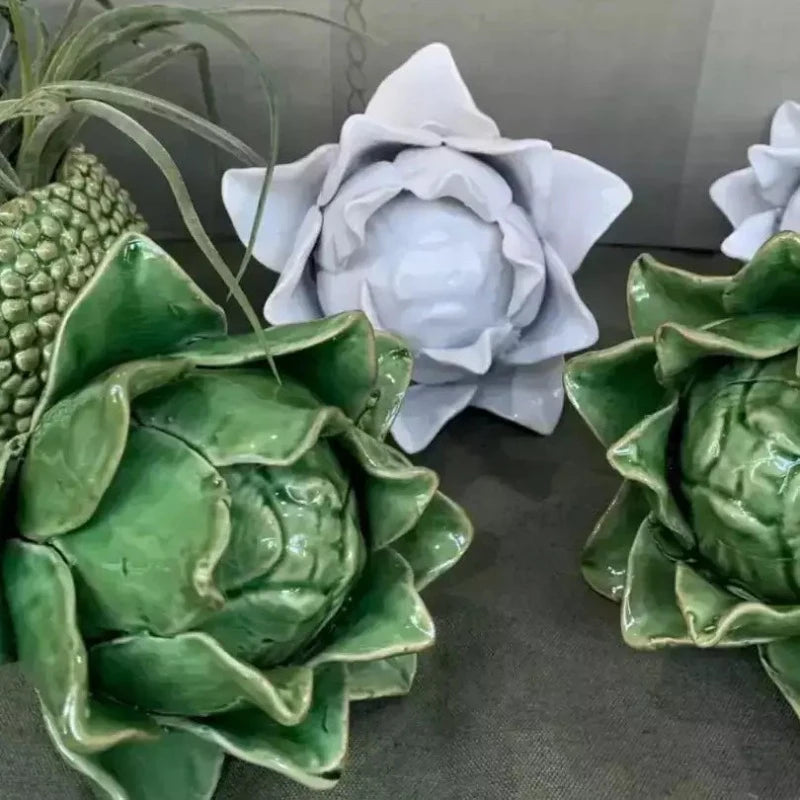 Artichoke - Green by Mediterranean Markets features green ceramic artichokes with a Sage Green glaze and glossy finish, contrasting with white matte ceramic flowers. Both rest on a gray surface, offering an artistic and decorative arrangement.