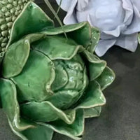 Thumbnail for A close-up of the Artichoke - Green by Mediterranean Markets shows its glossy Sage Green ceramic petals catching the light on a dark surface, with a partially visible white ceramic figure in the background adding contrast.