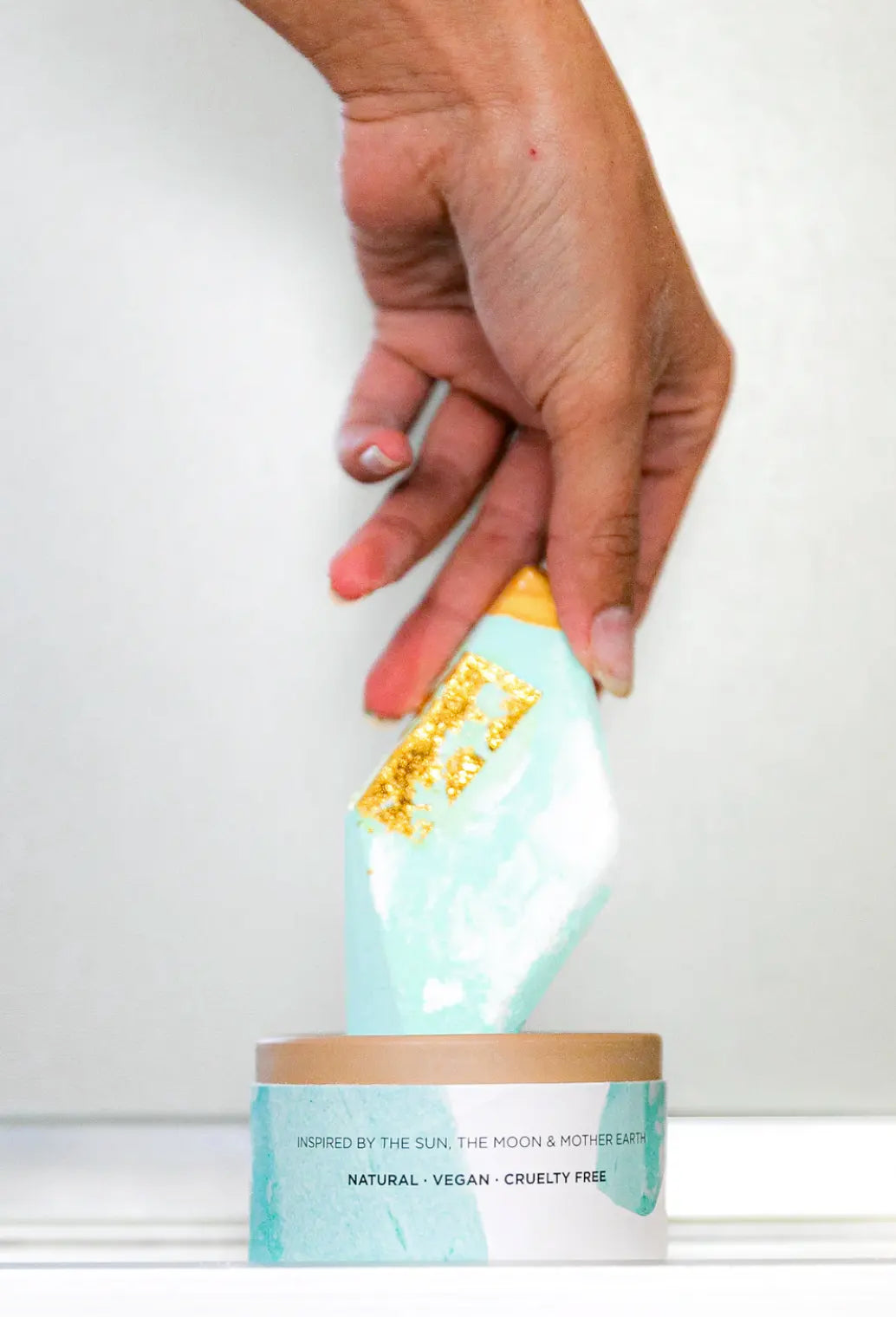 A hand is placing a Summer Salt Body Aquamarine Lemongrass Crystal Bath Bomb, infused with aromatherapy scents, into a box.