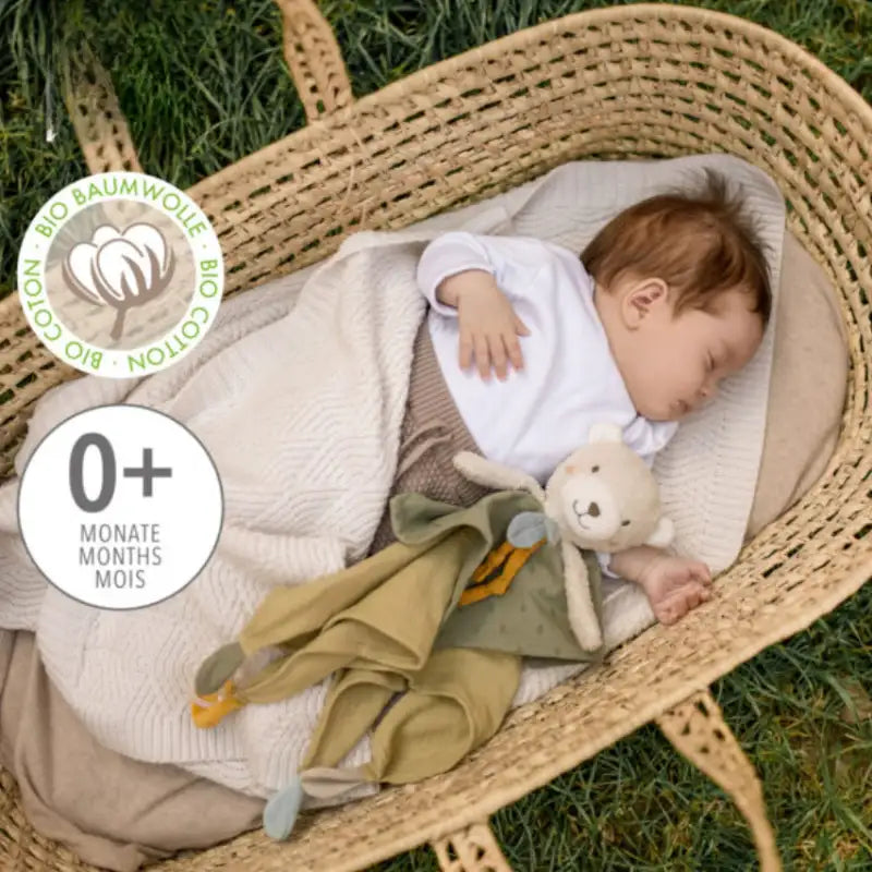 A baby naps in a woven basket on grass, cuddling a Pilbeam FehnNATUR Comforter Bear made with organic cotton, perfect for sensory development. The image includes a 0+ months label and Bio Cotton logo.