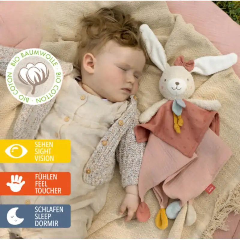 A baby sleeps on a pink blanket with the Pilbeam FehnNATUR Comforter Hare, ideal for sensory development. Icons show sight, touch, and sleep benefits. A badge in German and English confirms it’s made from organic cotton.