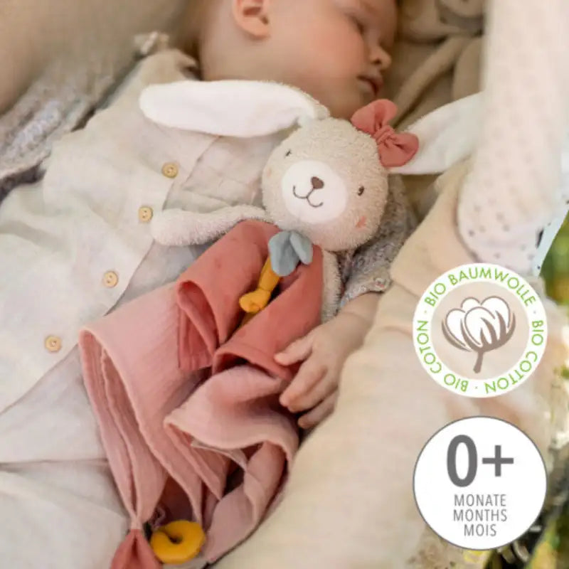 A baby cuddles the FehnNATUR Comforter Hare by Pilbeam, a soft white plush toy with pink dress and bow. The BIO COTTON badge shows it’s organic cotton, and the 0+ months label supports gentle sensory development.