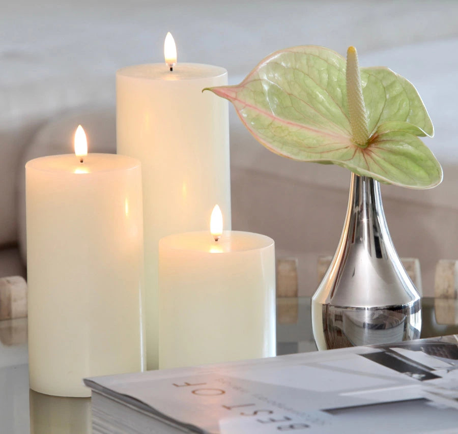 Three Enjoy Living Nordic White flameless LED candles on a table next to a magazine create a cozy ambiance lighting.