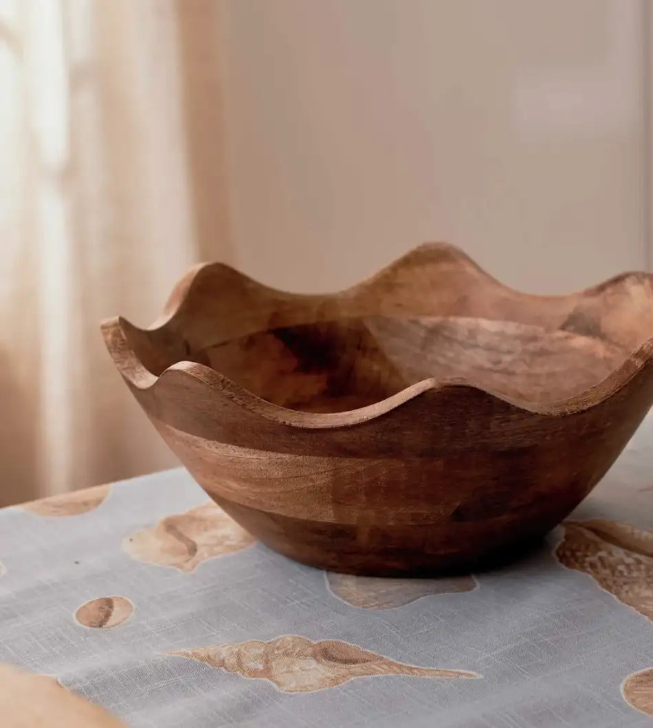 The Hazel Wavy Bowl by Madras Link, handcrafted from mango wood, rests on a light blue tablecloth with seashell accents—an ideal touch for contemporary dining spaces.