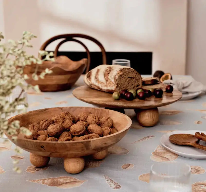 A dining table with a patterned tablecloth displays the Madras Link Hazel Wavy Bowl and wooden stands with walnuts, bread, cherries, and olives. Mango wood bowls and utensils add rustic charm to this modern dining scene.