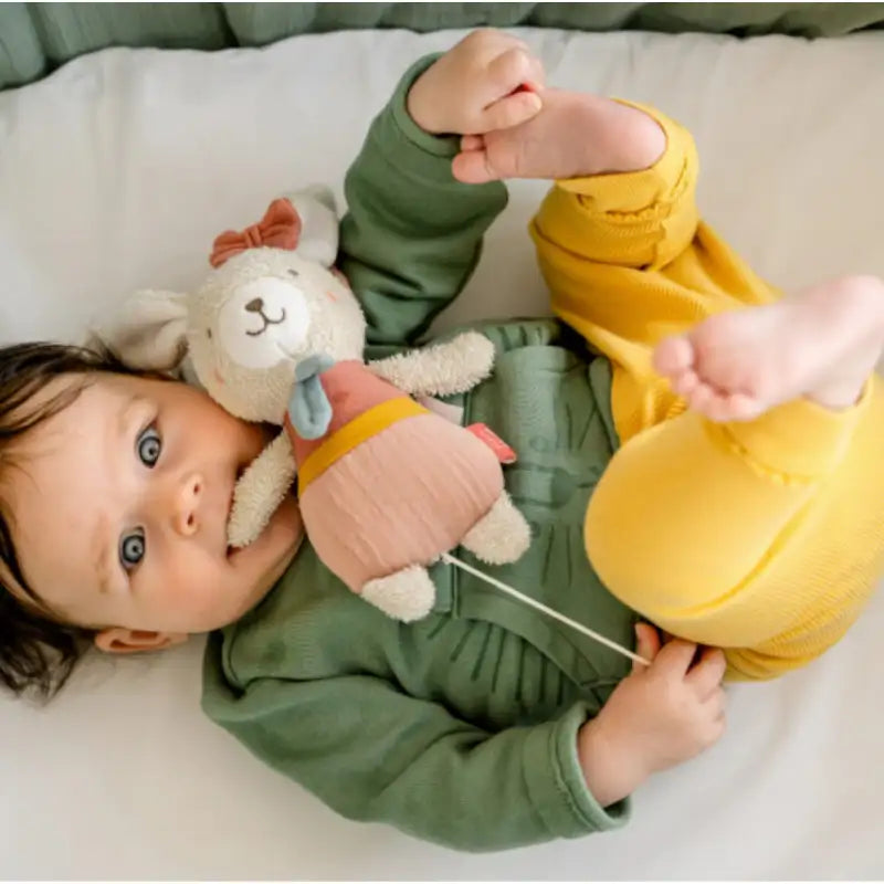 A baby with brown hair, dressed in a green organic cotton top and yellow pants, lies on a bed holding the Pilbeam FehnNATUR Mini Musical Bear. The baby gazes upward with wide blue eyes and bare feet raised.