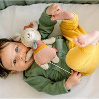 Thumbnail for A baby with brown hair, dressed in a green organic cotton top and yellow pants, lies on a bed holding the Pilbeam FehnNATUR Mini Musical Bear. The baby gazes upward with wide blue eyes and bare feet raised.