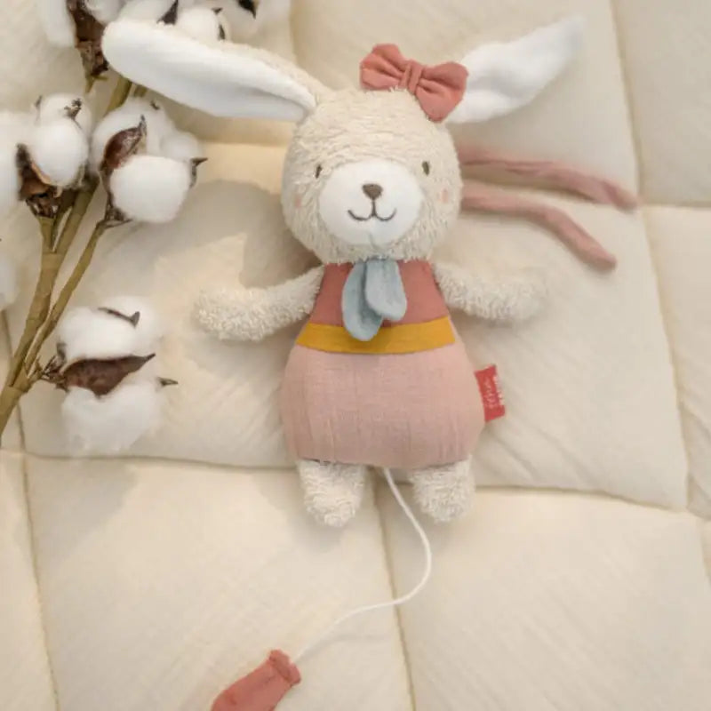 The FehnNATUR Mini Musical Hare by Pilbeam, made from organic cotton and dressed in a pink dress with red bow and blue scarf, rests on a quilted cream surface beside a cotton branch—a charming sleeping aid with pull cord.