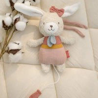 Thumbnail for The FehnNATUR Mini Musical Hare by Pilbeam, made from organic cotton and dressed in a pink dress with red bow and blue scarf, rests on a quilted cream surface beside a cotton branch—a charming sleeping aid with pull cord.