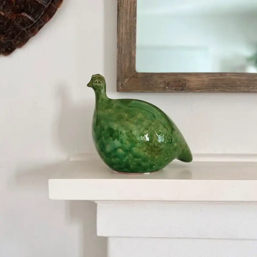 A Mode Guinea Fowl ceramic bird figurine by Mediterranean Markets rests on a white mantel beneath a wooden-framed mirror, with part of a wall decoration visible in the corner.