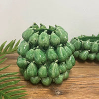 Thumbnail for The Mode Pear Vase by Mediterranean Markets is a green ceramic vase with glossy, pear-shaped clusters, inspired by classic Sicilian vases. A fern leaf sits in front on a wooden surface; another similar vase is partly visible behind.