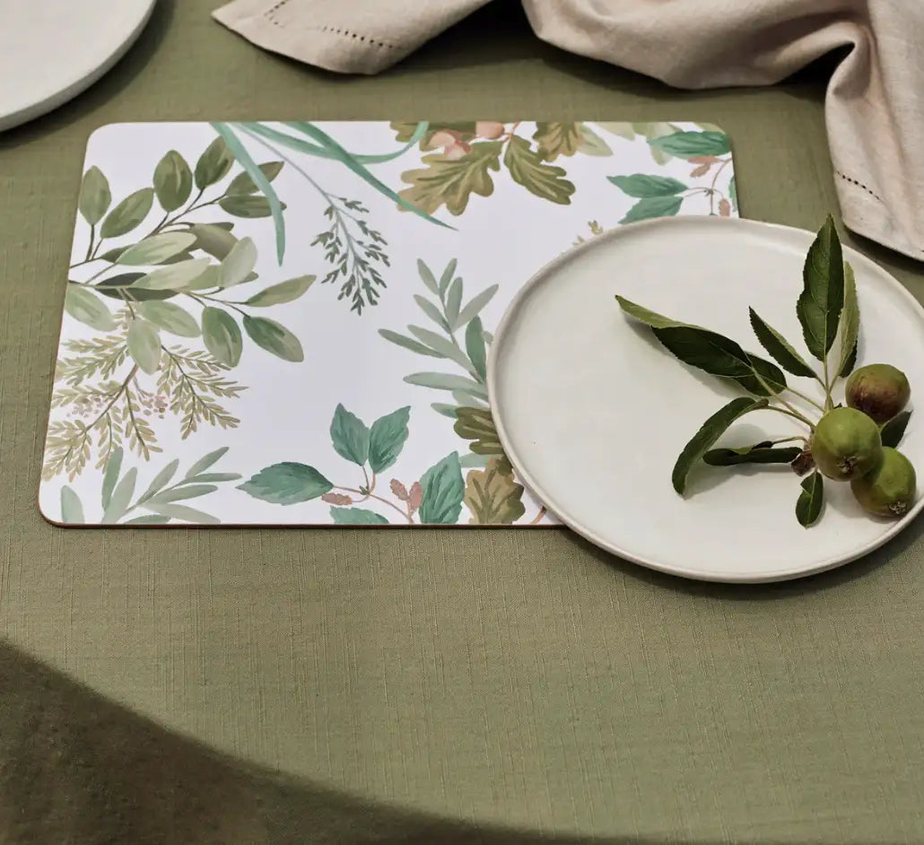 A white plate with green leaves and small fruits sits on a green tablecloth beside a Madras Link Placemat Naree Green Rectangle (Set of 4). Beige napkins are visible in the background.