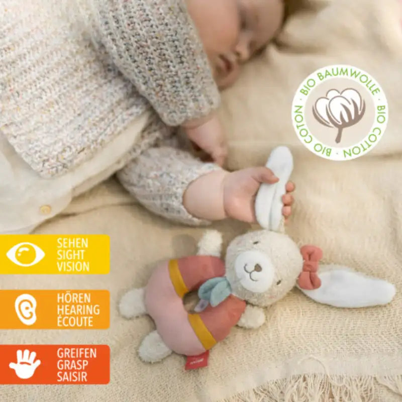 A baby lies on a beige blanket, reaching for the Pilbeam FehnNATUR Soft Ring Rattle Hare. Icons highlight senses: Sight, Hearing, Grasp. A circular label reads “Bio Cotton/Bio-Baumwolle,” emphasizing organic cotton and development.