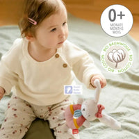Thumbnail for A baby in a cream sweater and patterned pants sits on a soft green blanket holding the Pilbeam FehnNATUR Soft Ring Rattle Hare, an organic cotton plush toy suitable for ages 0+ months and perfect for encouraging motor skill development.