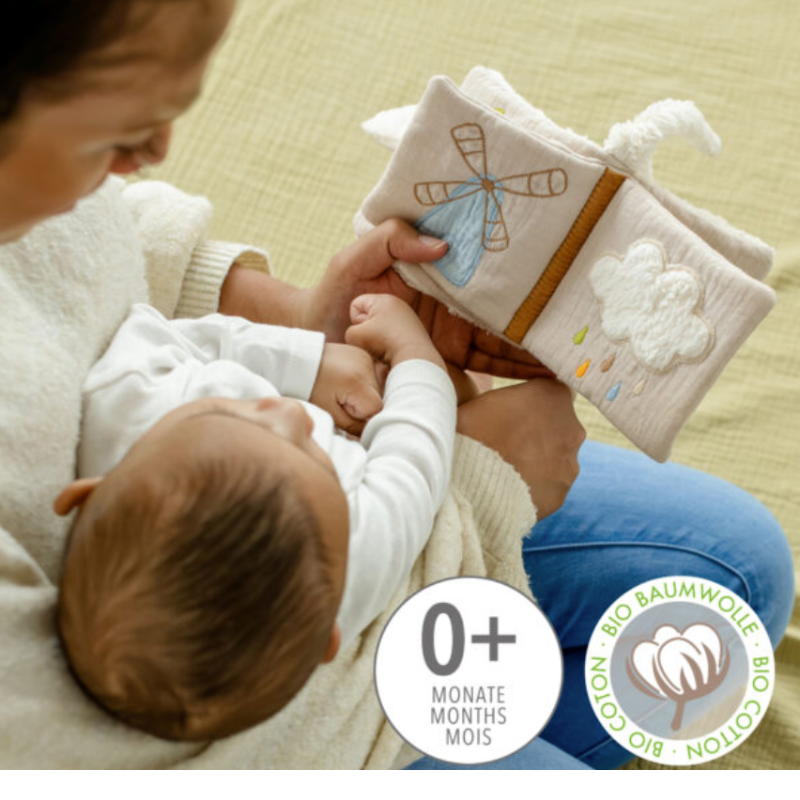 An adult shows a baby the FehnNATTUR Soft Book Sheep by Pilbeam. Made from organic cotton, this sensory toy features embroidered windmill and cloud images. A badge marks it as suitable for ages 0+, making it a perfect baby gift.