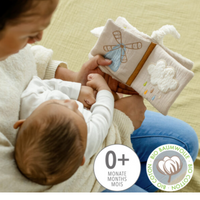 Thumbnail for An adult shows a baby the FehnNATTUR Soft Book Sheep by Pilbeam. Made from organic cotton, this sensory toy features embroidered windmill and cloud images. A badge marks it as suitable for ages 0+, making it a perfect baby gift.