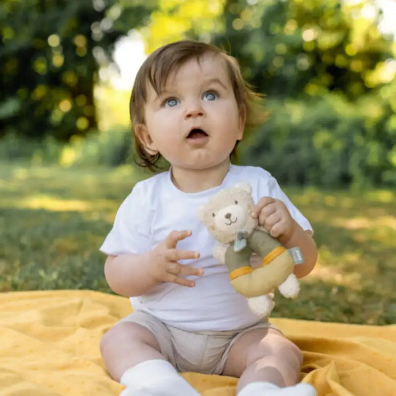A baby with light brown hair and blue eyes sits outdoors on a yellow blanket, holding the Pilbeam FehnNATUR Soft Ring Rattle Bear—an organic cotton toy perfect for motor skill development—surrounded by green trees and grass.