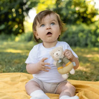 Thumbnail for A baby with light brown hair and blue eyes sits outdoors on a yellow blanket, holding the Pilbeam FehnNATUR Soft Ring Rattle Bear—an organic cotton toy perfect for motor skill development—surrounded by green trees and grass.