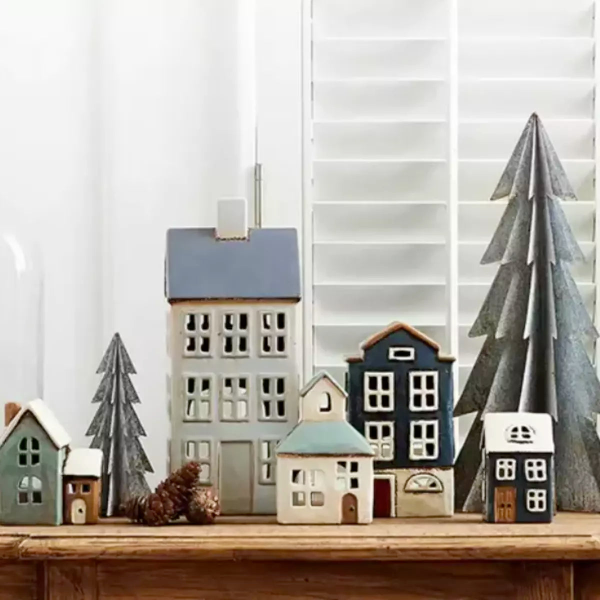 French Country Collections' A Christmas Village Mini Church tea light holder mimics a charming Christmas Village, with small wooden houses nestled on a mantle alongside pine trees.