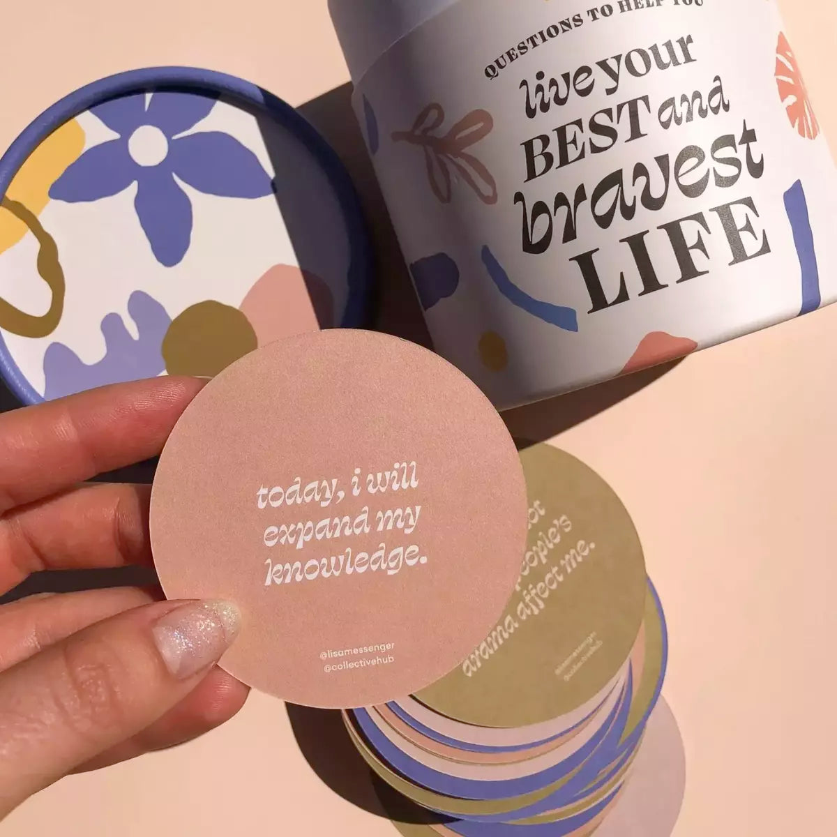 A person holding a tin from Collective Hub with a card that reads, 'Questions to Help You Live Your Best and Bravest Life' in an abstract floral tube lid box design.